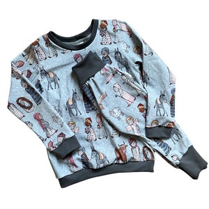 May include: A blue children's pajama set with a long-sleeved top and pants. The fabric features a repeating pattern of a nativity scene with figures like Mary, Joseph, and a donkey.