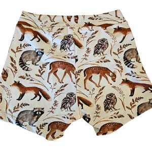 Op de afbeelding: Witte shorts met een herhaald patroon van bosdieren, waaronder vossen, wasberen, herten en uilen. De shorts hebben een comfortabele pasvorm en zijn gemaakt van een zacht materiaal. Het ontwerp gebruikt bruine en beige kleuren.