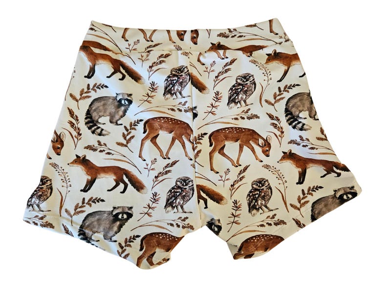 Op de afbeelding: Witte shorts met een herhaald patroon van bosdieren, waaronder vossen, wasberen, herten en uilen. De shorts hebben een comfortabele pasvorm en zijn gemaakt van een zacht materiaal. Het ontwerp gebruikt bruine en beige kleuren.