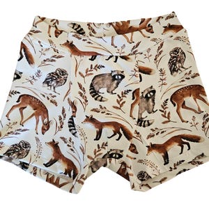 Op de afbeelding: Cr&egrave;mekleurige shorts met een herhaald patroon van bosdieren, waaronder vossen, herten, wasberen en uilen. De shorts hebben een hoge tailleband en zijn versierd met bruin gebladerte.
