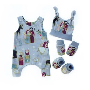 Könnte beinhalten: Ein Baby-Outfit-Set mit einem blauen Strampler, einer Mütze, Stiefeln und Fäustlingen. Der Stoff hat einen Weihnachts-Krippen-Druck mit goldenen Akzenten.