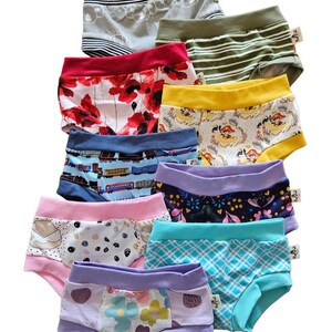 Pode incluir: Uma cole&ccedil;&atilde;o de nove pares de roupa interior infantil com v&aacute;rios padr&otilde;es, incluindo coalas, riscas verdes, papoilas vermelhas, faixas amarelas, comboios, motivos florais, pintas, xadrez azul e flores coloridas, cada uma com uma pequena etiqueta.