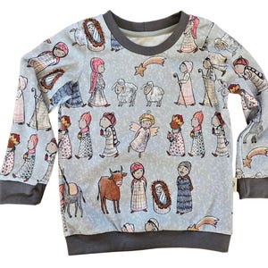 May include: A light blue long-sleeved sweatshirt featuring a repeating pattern of nativity figures, angels, and animals. The design includes a dark gray crew neck and cuffs. The illustrations are in soft colors, creating a whimsical and festive look.