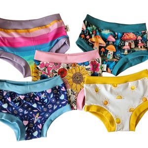 Pode incluir: Cinco pares de roupa interior de cintura subida com v&aacute;rios padr&otilde;es: um com riscas horizontais arco-&iacute;ris, um com cogumelos, um com girass&oacute;is e abelhas, um com design floral azul escuro e um com s&oacute;is amarelos sobre fundo branco.