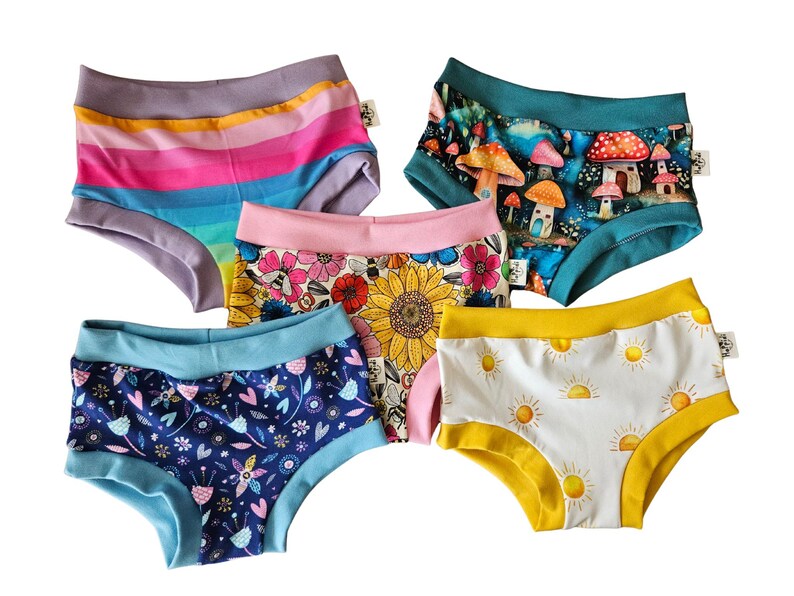 Pode incluir: Cinco pares de roupa interior de cintura subida com v&aacute;rios padr&otilde;es: um com riscas horizontais arco-&iacute;ris, um com cogumelos, um com girass&oacute;is e abelhas, um com design floral azul escuro e um com s&oacute;is amarelos sobre fundo branco.