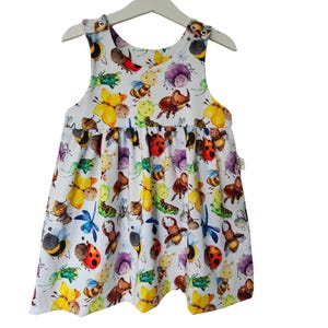 Puede incluir: Un vestido blanco sin mangas con un estampado colorido de varios insectos, incluyendo mariquitas, mariposas y abejas.