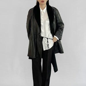 May include: A black leather coat with a black fur collar, worn over a white button-down shirt and black pants. The coat has a long, flowing design and a matching black belt. The outfit is completed with black shoes.