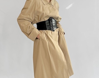 May include: A tan trench coat with a black corset belt. The coat has a collared neckline, long sleeves, and pockets. The black corset belt cinches the waist. The model is wearing black flats and sheer black stockings.