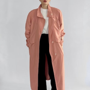 Puede incluir: Un abrigo largo tipo gabardina de color coral con diseño de doble botonadura. El abrigo presenta un cuello con solapas, cierres de botones y un cinturón a juego. El modelo lleva una camiseta blanca y pantalones negros.