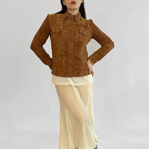 May include: A brown suede button-up shirt with two chest pockets. The shirt is paired with a cream-colored top and a long, flowing cream skirt. Brown leather boots complete the outfit. The outfit is displayed against a white background.