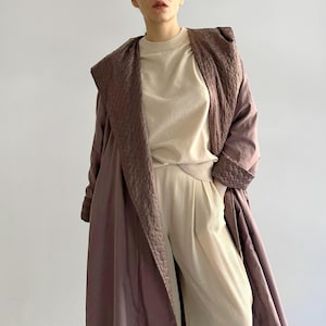 Può includere: Un lungo cappotto color malva con cappuccio e fodera trapuntata. Il cappotto è aperto e il cappuccio è abbassato. La persona che indossa il cappotto indossa una maglia e pantaloni color crema.
