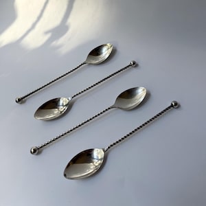 May include: Four silver spoons with twisted handles and a small ball at the end. The spoons have an oval bowl shape. The spoons are arranged diagonally on a white surface, with a shadow in the background.