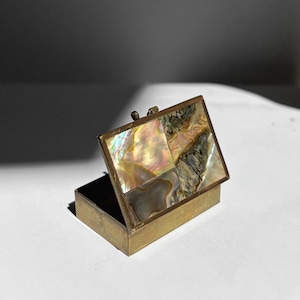 May include: A rectangular brass box with a hinged lid featuring an inlaid mosaic of iridescent mother-of-pearl and abalone shell segments. The metal frame has a muted, aged finish, and the lid is propped open to reveal the interior structure.