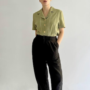 May include: A light green short-sleeved button-up shirt with white stitching and black high-waisted pants. The person is wearing white and black sneakers.