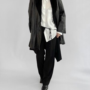 May include: A black leather coat with a faux fur collar, worn over a white button-down shirt and black pants. The coat has a long, flowing design. The outfit is completed with black shoes. The overall style is minimalist and chic.