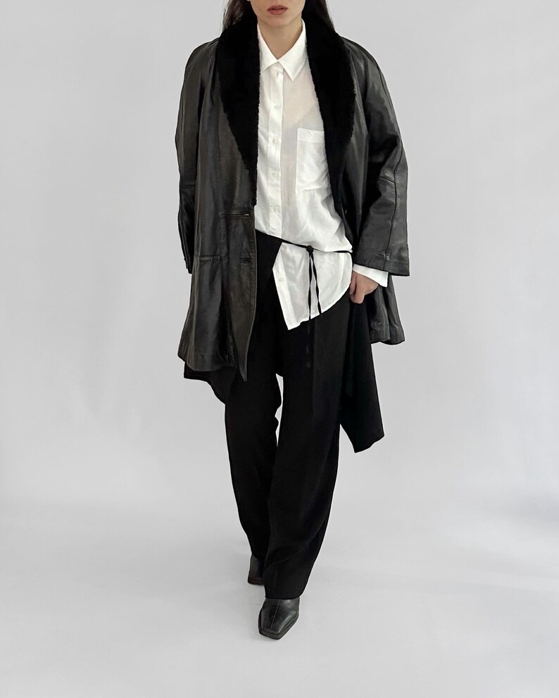 May include: A black leather coat with a faux fur collar, worn over a white button-down shirt and black pants. The coat has a long, flowing design. The outfit is completed with black shoes. The overall style is minimalist and chic.
