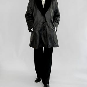 May include: A black leather coat with a black faux fur collar and a button-down front. The coat has two front pockets and is paired with black pants and shoes. The outfit is set against a plain white background.