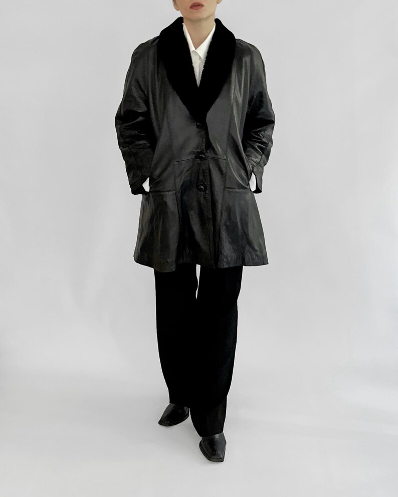 May include: A black leather coat with a black faux fur collar and a button-down front. The coat has two front pockets and is paired with black pants and shoes. The outfit is set against a plain white background.