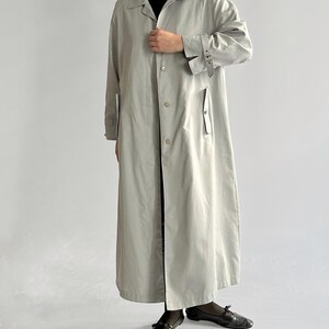 May include: A long, light gray trench coat with a belt and button closure. The coat has a collar and long sleeves.