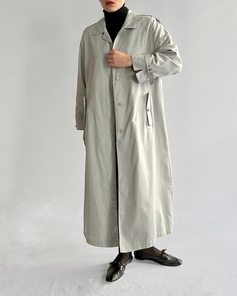 May include: A long, light gray trench coat with a belt and button closure. The coat has a collar and long sleeves.