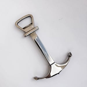 Könnte beinhalten: Ein silberfarbener Flaschenöffner aus Metall in Form eines nautischen Ankers. Das Oberteil verfügt über eine rechteckige Öffnung zum Abhebeln von Kronkorken, während das Unterteil aus einem geschwungenen Ankerdesign mit Spitzen besteht.
