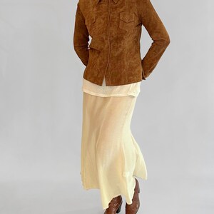 May include: A brown suede jacket with a collared neckline and two chest pockets. The jacket is worn over a cream-colored top and a long, flowing, pale yellow skirt. Brown leather boots complete the outfit.
