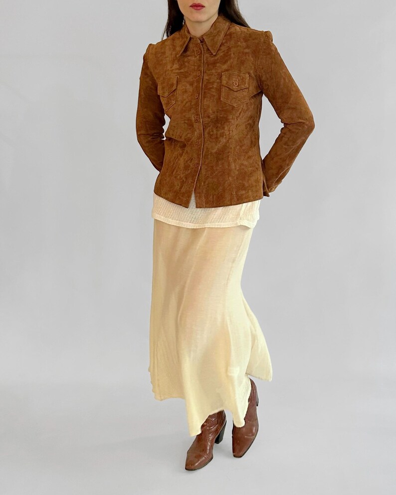 May include: A brown suede jacket with a collared neckline and two chest pockets. The jacket is worn over a cream-colored top and a long, flowing, pale yellow skirt. Brown leather boots complete the outfit.