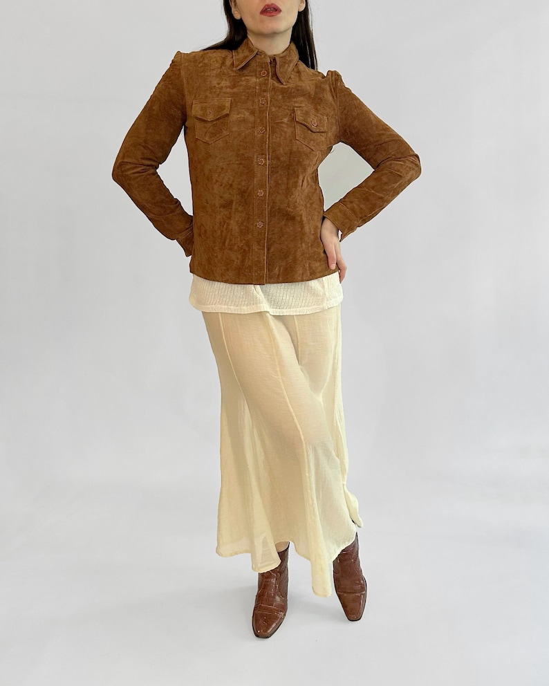 May include: A brown suede button-up shirt with two chest pockets. The shirt is paired with a cream-colored top and a long, flowing cream skirt. Brown leather boots complete the outfit. The outfit is displayed against a white background.
