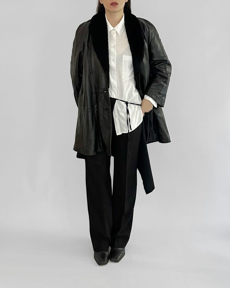 May include: A black leather coat with a black fur collar, worn over a white button-down shirt and black pants. The coat has a long, flowing design and a matching black belt. The outfit is completed with black shoes.