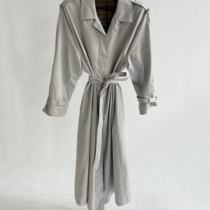 May include: A light gray trench coat with a plaid lining and a belt. The coat has long sleeves and a collar.