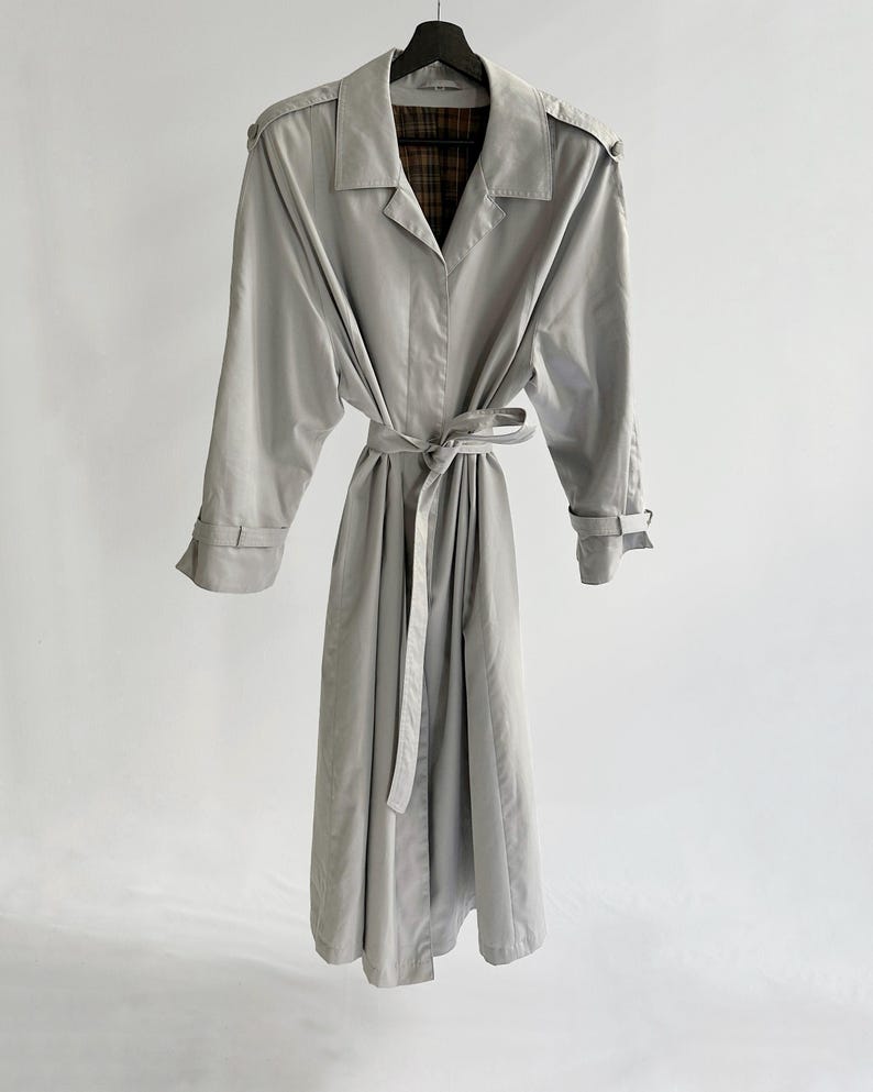 May include: A light gray trench coat with a plaid lining and a belt. The coat has long sleeves and a collar.