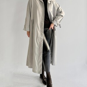 May include: A long, light gray trench coat with a belt and a button closure. The coat is worn over a black turtleneck and black pants.