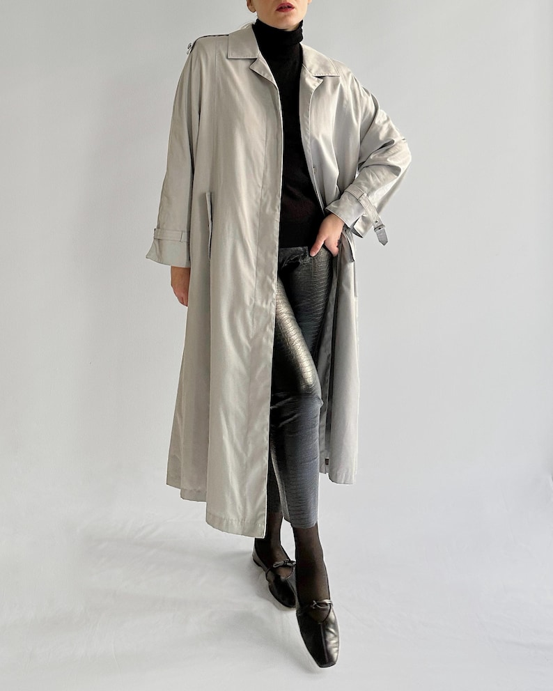 May include: A long, light gray trench coat with a belt and a button closure. The coat is worn over a black turtleneck and black pants.