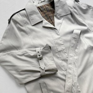 May include: A light gray trench coat with a plaid lining and a belt. The coat has a button closure and a single button on the pocket flap.