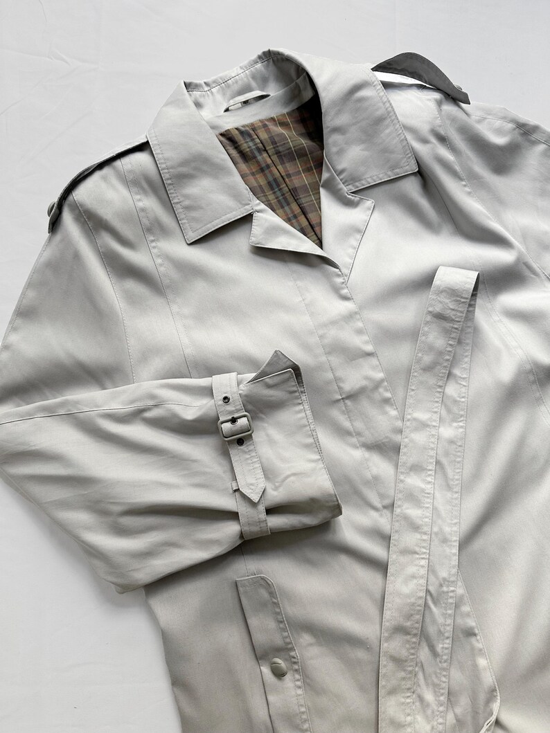 May include: A light gray trench coat with a plaid lining and a belt. The coat has a button closure and a single button on the pocket flap.