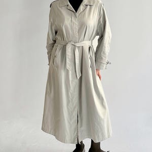 May include: A long, light gray trench coat with a belt and a button closure. The coat has a classic design and is made of a lightweight fabric.
