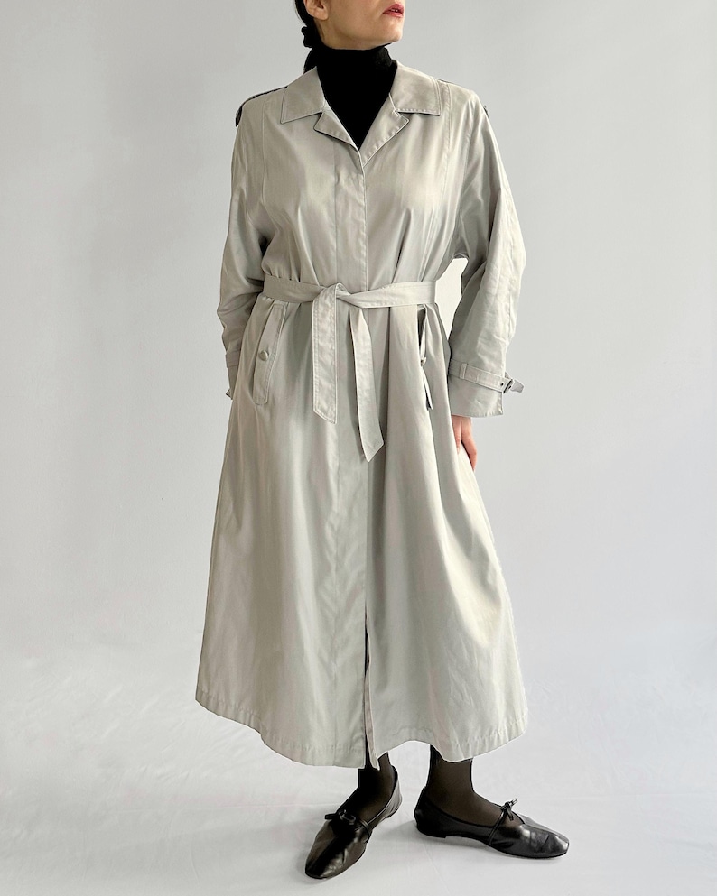 May include: A long, light gray trench coat with a belt and a button closure. The coat has a classic design and is made of a lightweight fabric.