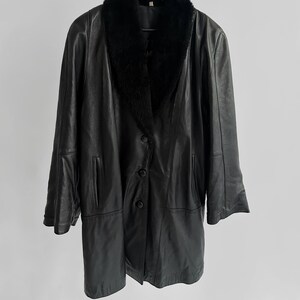 May include: A black leather coat with a faux fur collar hangs from a black hanger. The coat has a button-down front, two front pockets, and long sleeves. The coat is a classic style, suitable for cold weather.
