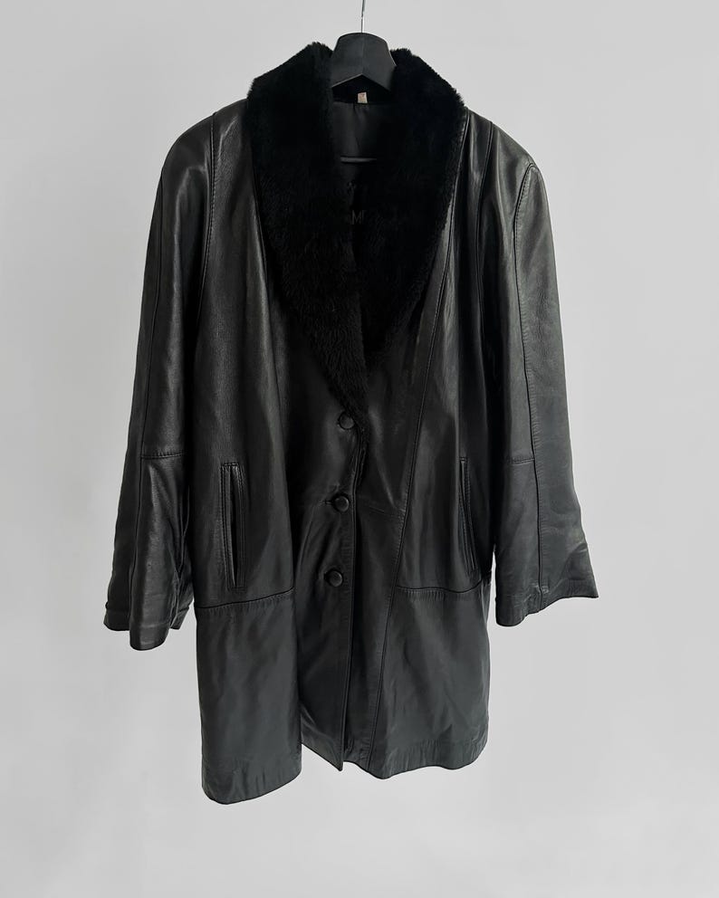May include: A black leather coat with a faux fur collar hangs from a black hanger. The coat has a button-down front, two front pockets, and long sleeves. The coat is a classic style, suitable for cold weather.