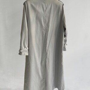 May include: A light gray trench coat with a long, flowing silhouette. The coat has a classic design with a double-breasted closure, a belt, and a collar.
