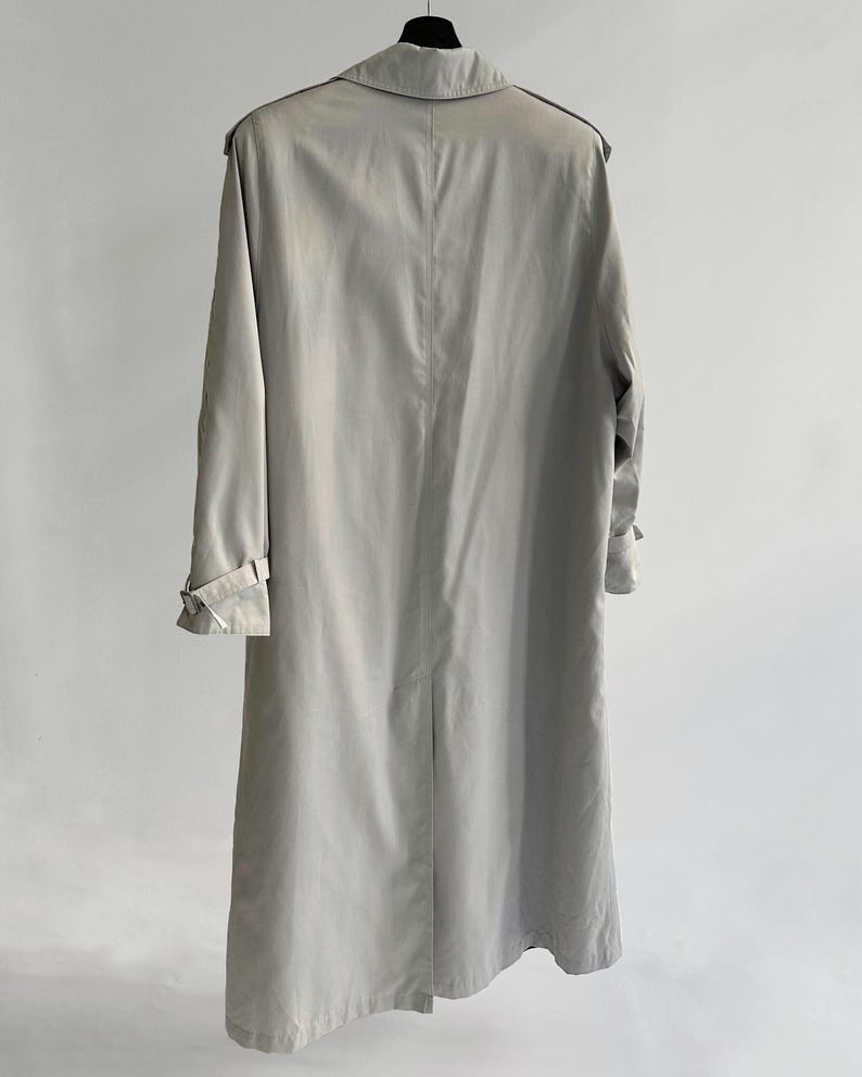 May include: A light gray trench coat with a long, flowing silhouette. The coat has a classic design with a double-breasted closure, a belt, and a collar.