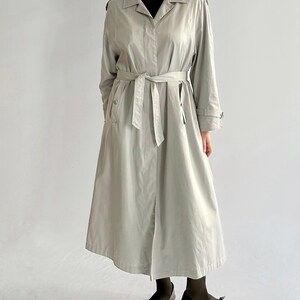 May include: A light gray trench coat with a black belt and black buttons. The coat has a long, flowing silhouette and a classic collar.
