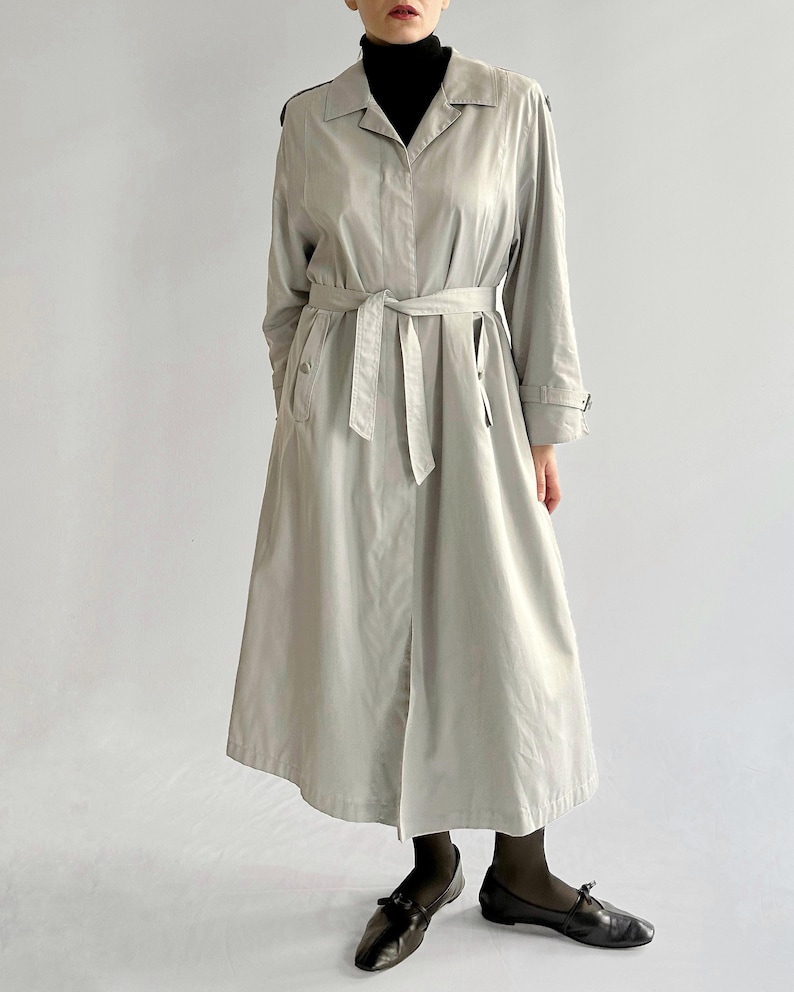 May include: A light gray trench coat with a black belt and black buttons. The coat has a long, flowing silhouette and a classic collar.