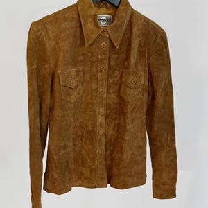 May include: A brown suede button-up shirt with a collared neckline and two chest pockets. The long-sleeved shirt is hanging on a black hanger against a white background. The shirt has a classic, vintage style.