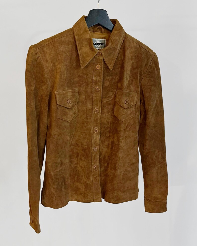 May include: A brown suede button-up shirt with a collared neckline and two chest pockets. The long-sleeved shirt is hanging on a black hanger against a white background. The shirt has a classic, vintage style.