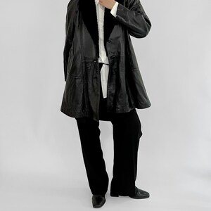 May include: A black leather coat with a black fur collar and a button-down front. The coat is paired with black pants and black shoes. The outfit is set against a plain white background.