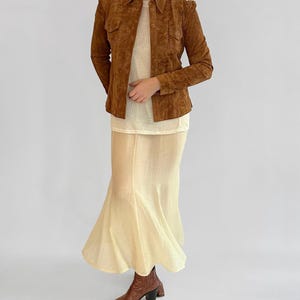 May include: A brown suede jacket with a collared neckline and button closures. The jacket is paired with a cream-colored top and a long, flowing skirt. Brown leather ankle boots complete the outfit, showcasing a vintage aesthetic.