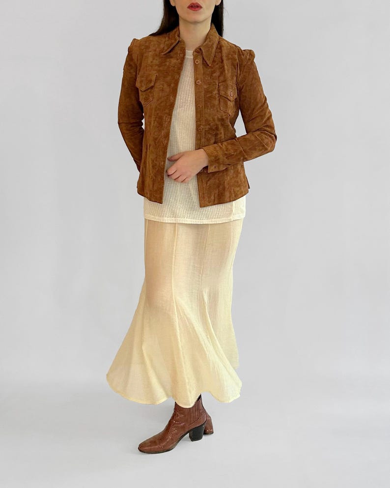 May include: A brown suede jacket with a collared neckline and button closures. The jacket is paired with a cream-colored top and a long, flowing skirt. Brown leather ankle boots complete the outfit, showcasing a vintage aesthetic.