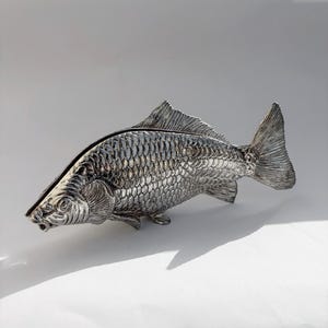 Pode incluir: Uma caixa de metal prateado em forma de peixe com tampa articulada. O exterior apresenta um padrão de escamas em relevo detalhado que cobre o corpo, as barbatanas e a cauda. O objeto tem um acabamento metálico com áreas escuras.