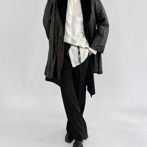 May include: A black leather coat with a faux fur collar, worn over a white button-down shirt and black pants. The coat has a long, flowing design. The outfit is completed with black shoes.